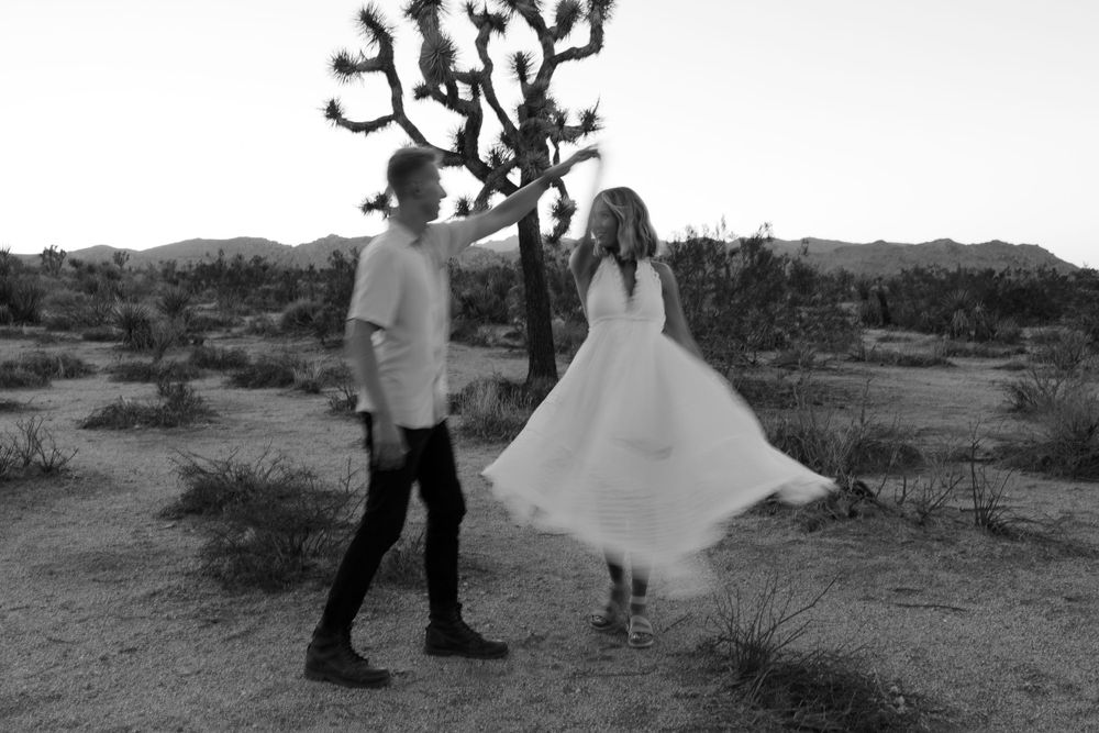 Couple poses in black and white desert landscape with Joshua tree silhouette at sunset.