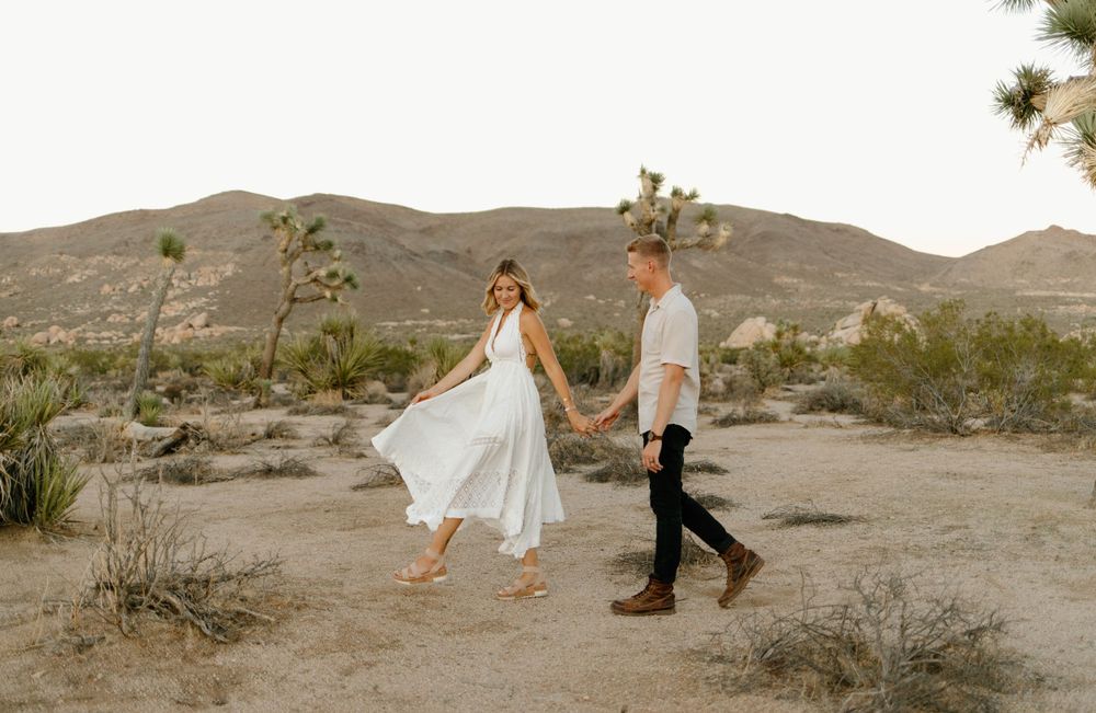 Couple walks hand in hand through desert landscape with mountains and Joshua trees in background.
