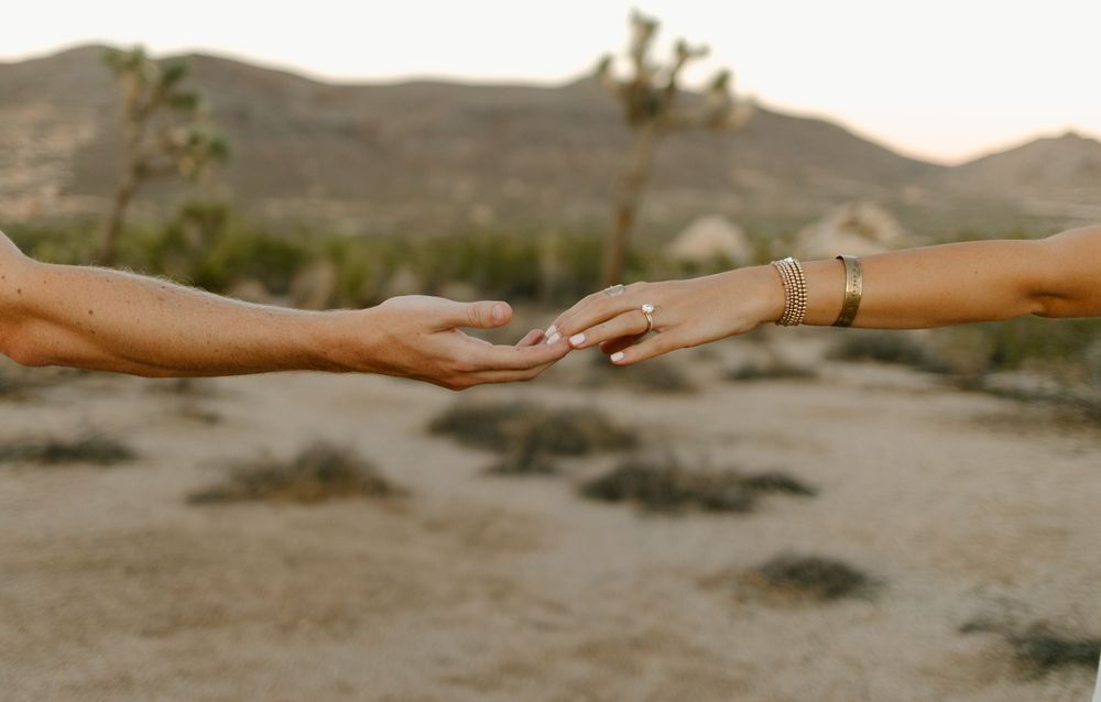 Close-up of hands reaching for each other against desert landscape backdrop.