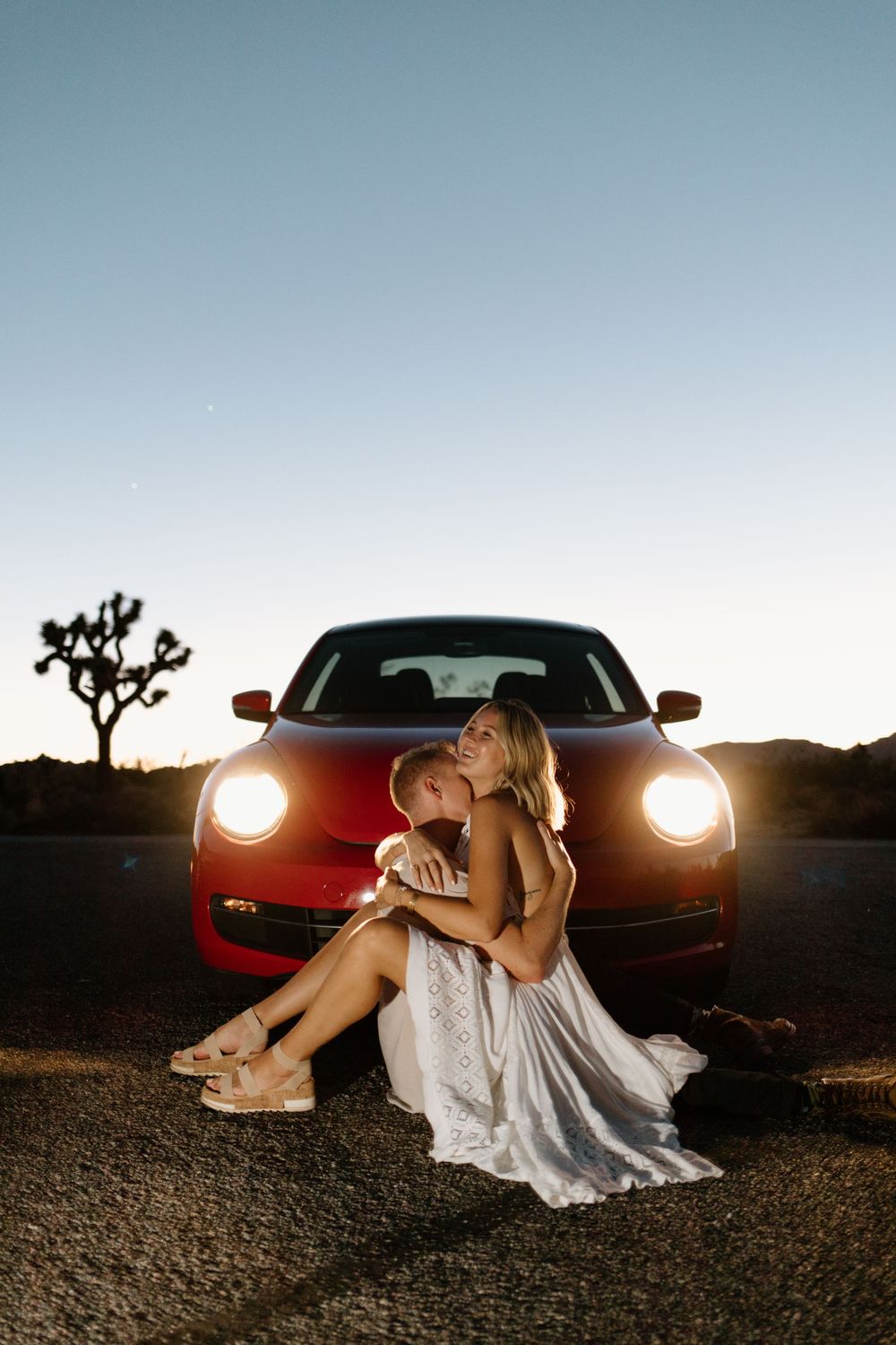 Woman in white dress poses by red car with headlights on at dusk in desert setting.