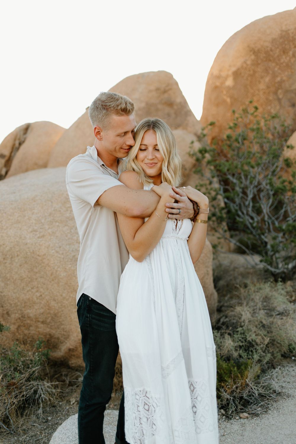 Couple embracing tenderly against backdrop of large desert boulders during golden hour.