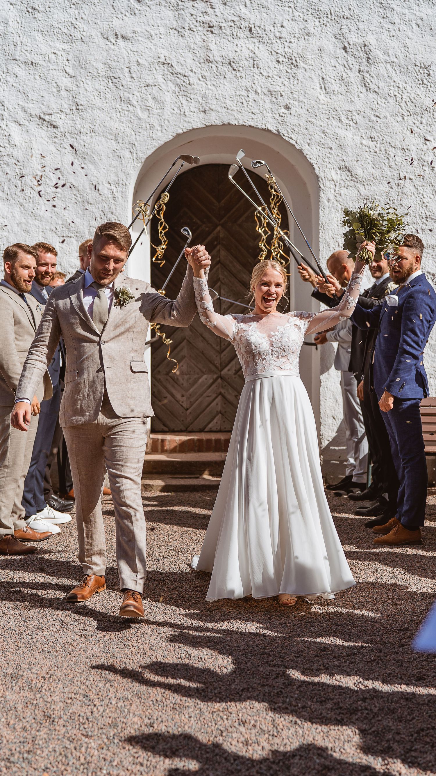Joyful newlyweds raise hands under golf club arch with groomsmen celebrating outside white church entrance.