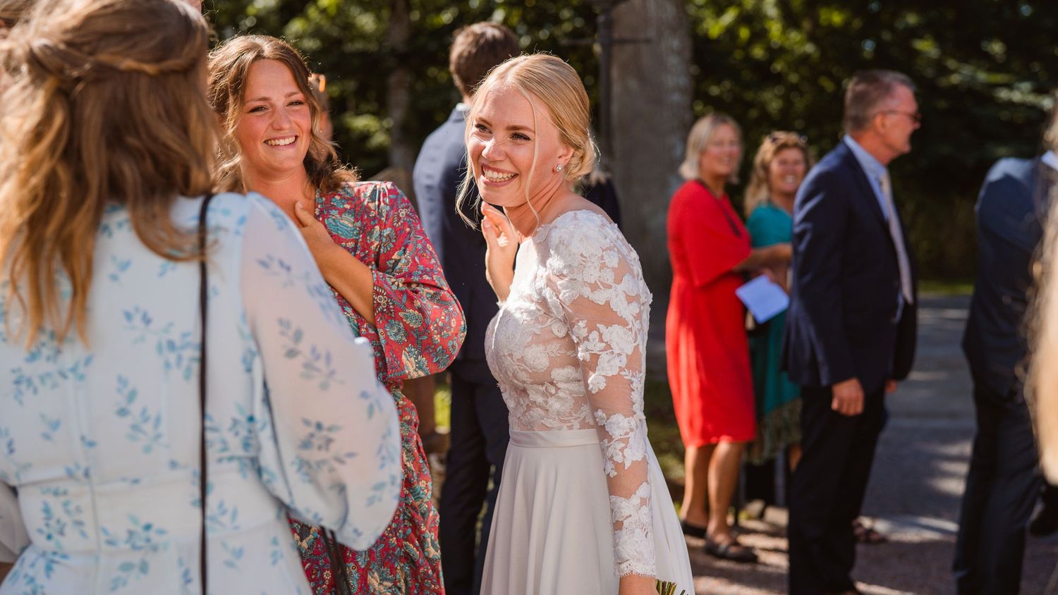 Laughing bride in lace gown greets smiling guests outdoors at sunny wedding reception gathering.