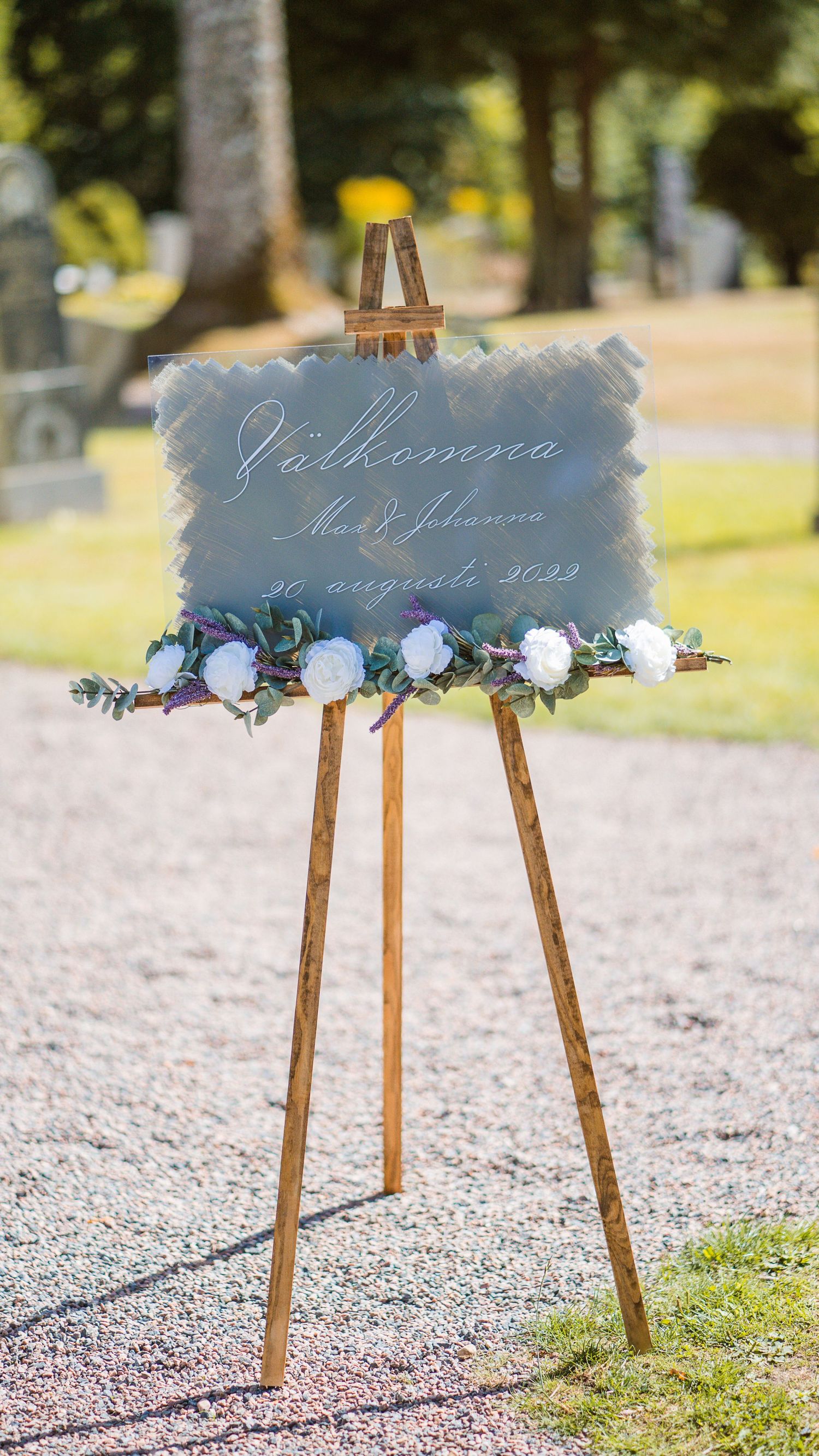 Elegant wedding welcome sign on wooden easel decorated with white roses and greenery.