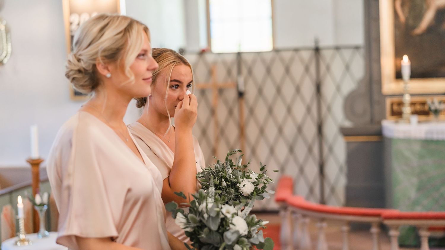 Two emotional bridesmaids in blush dresses watch wedding ceremony, one wiping tears while holding a bouquet.