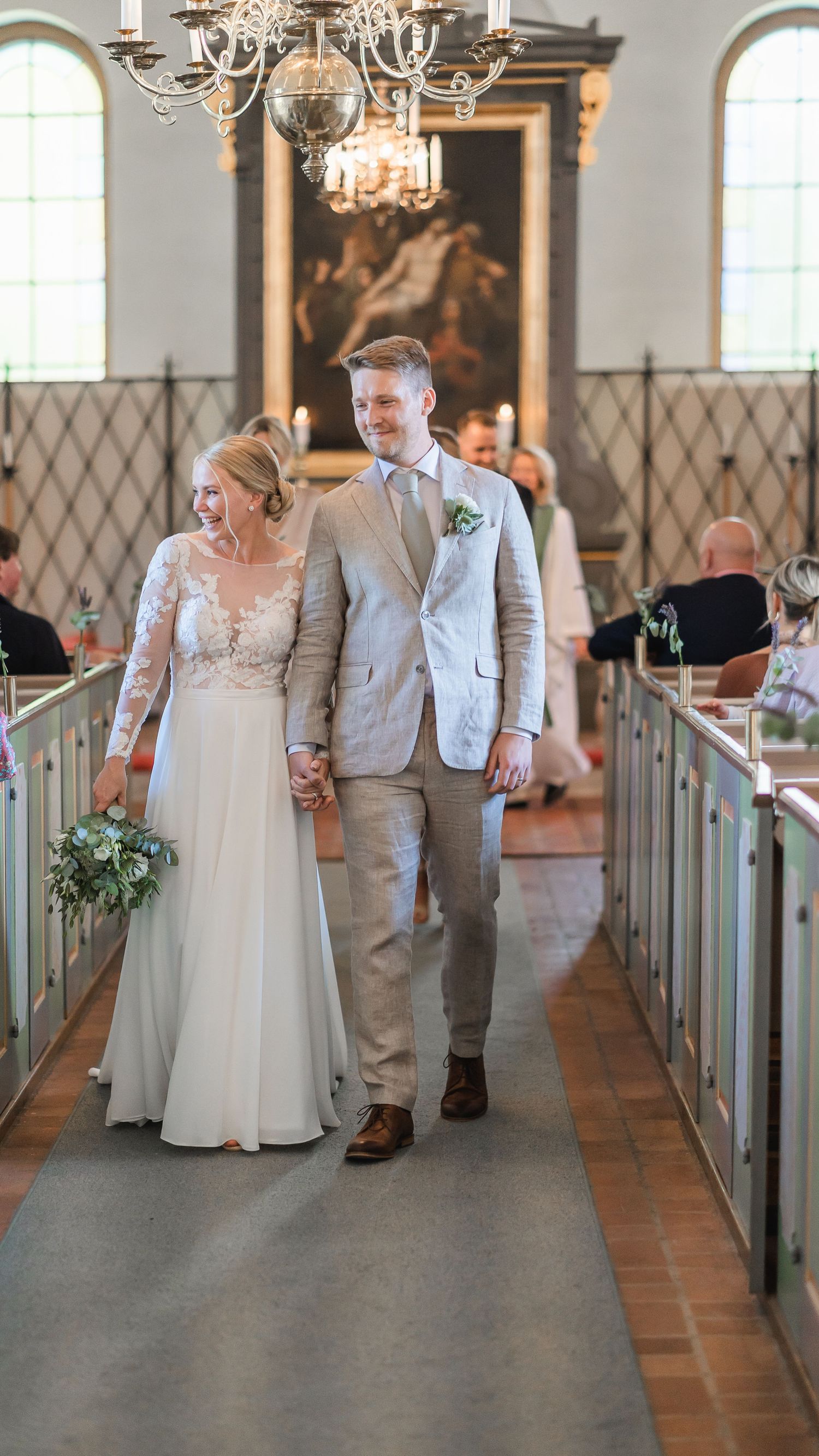 Smiling newlyweds walk down church aisle, bride holding greenery bouquet in white lace gown and groom in beige suit.