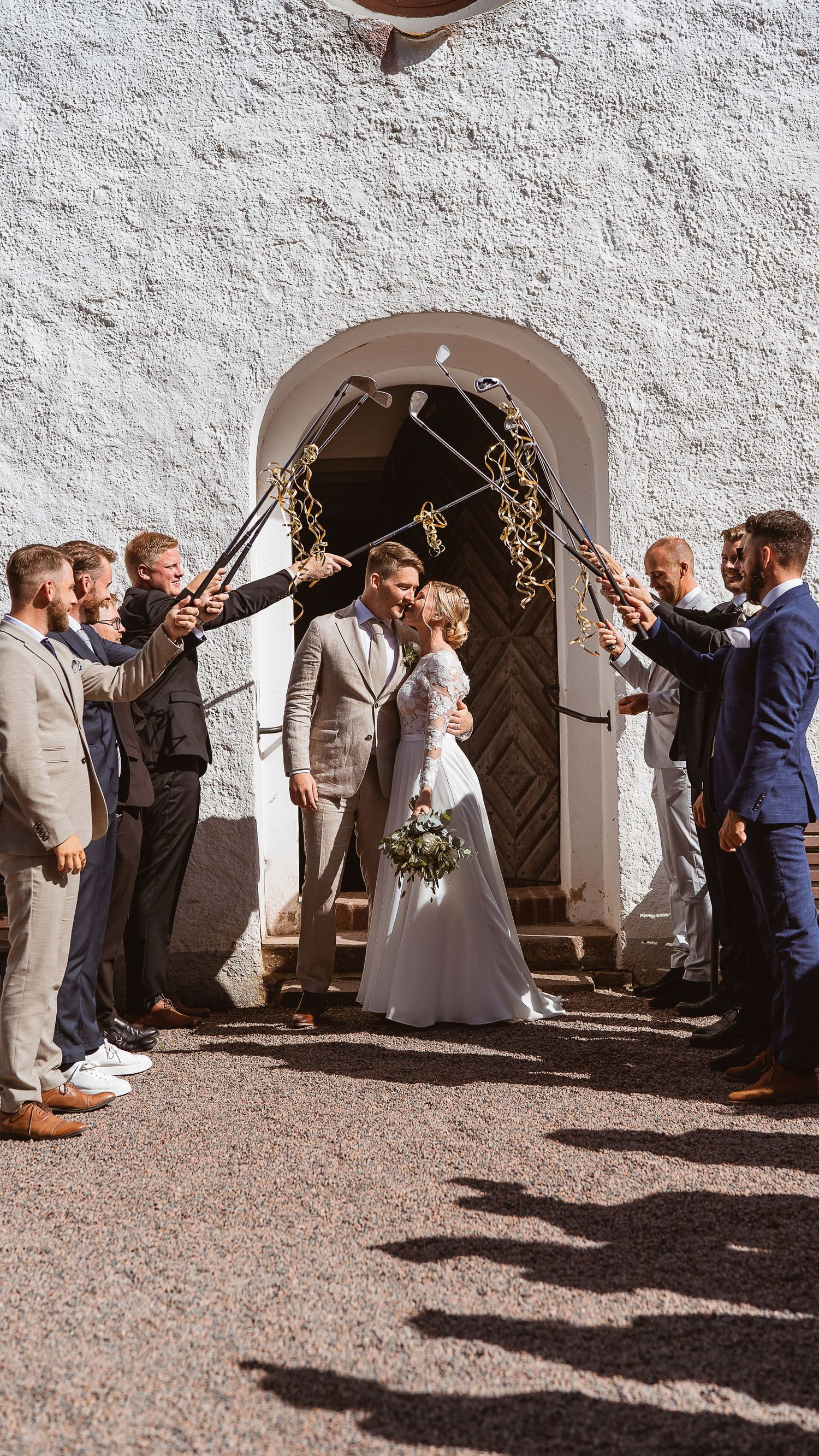 Newlyweds share a kiss under a golf club arch surrounded by groomsmen outside a white church.