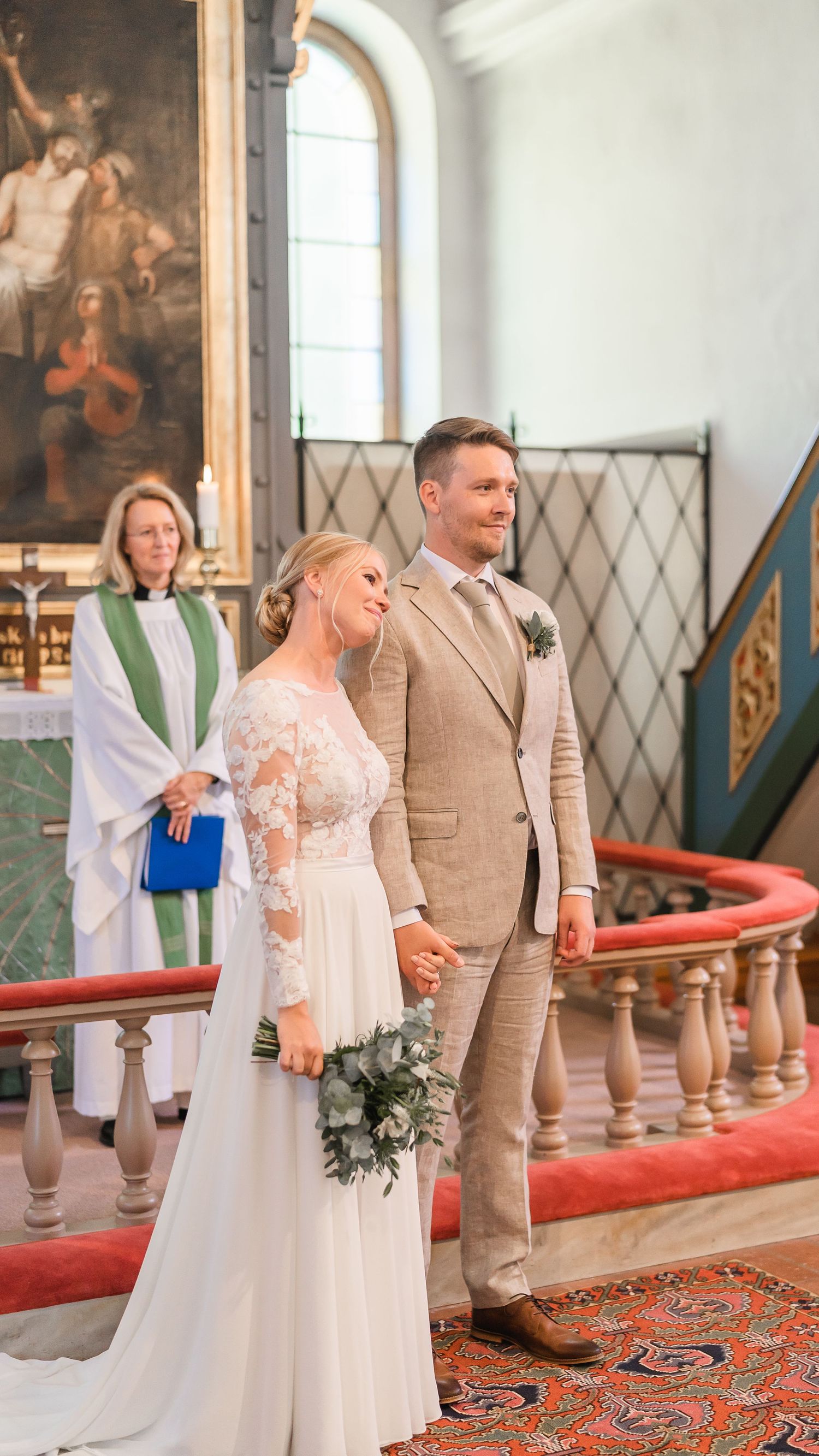 Bride in lace wedding dress and groom in tan suit hold hands at church altar with officiant behind them.