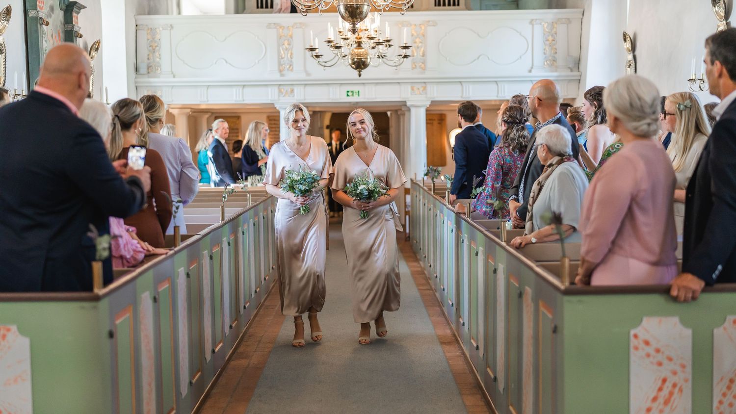 Two bridesmaids in champagne dresses walk down a church aisle carrying bouquets at a wedding ceremony.