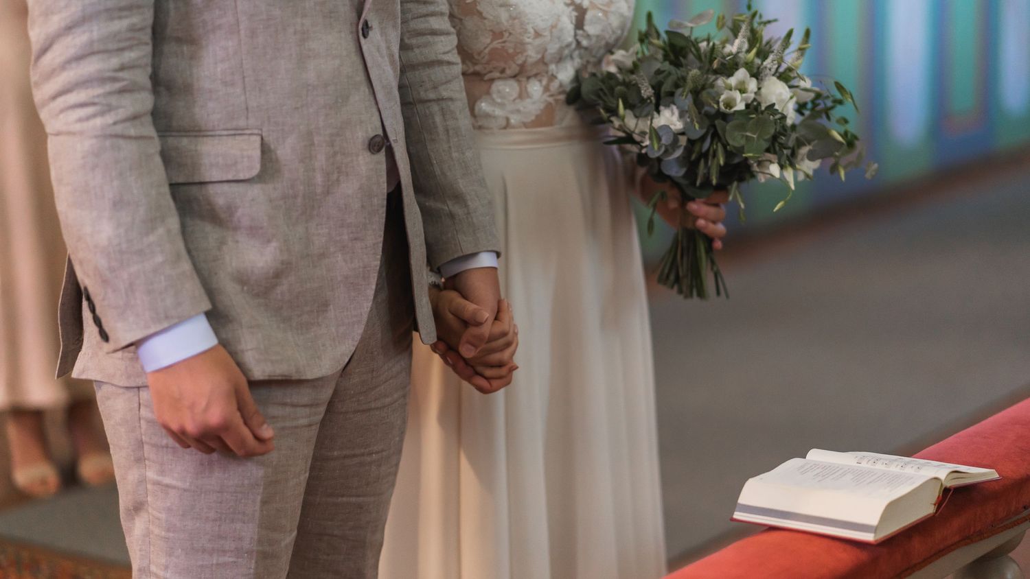 Bride and groom hold hands at the altar with a wedding bouquet and open Bible nearby.