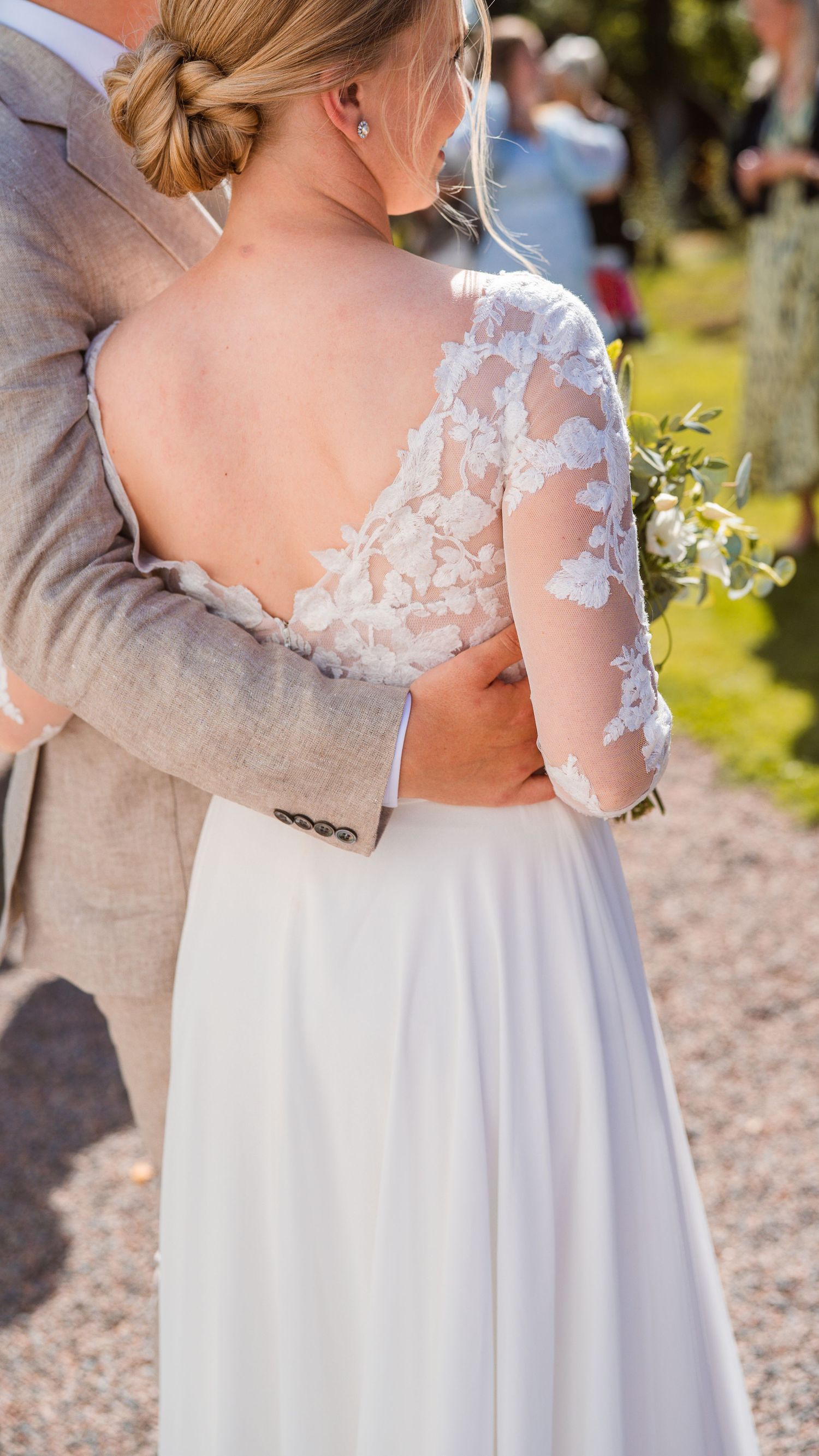 Close-up of bride's intricate lace back wedding gown as groom in gray suit embraces her outdoors.