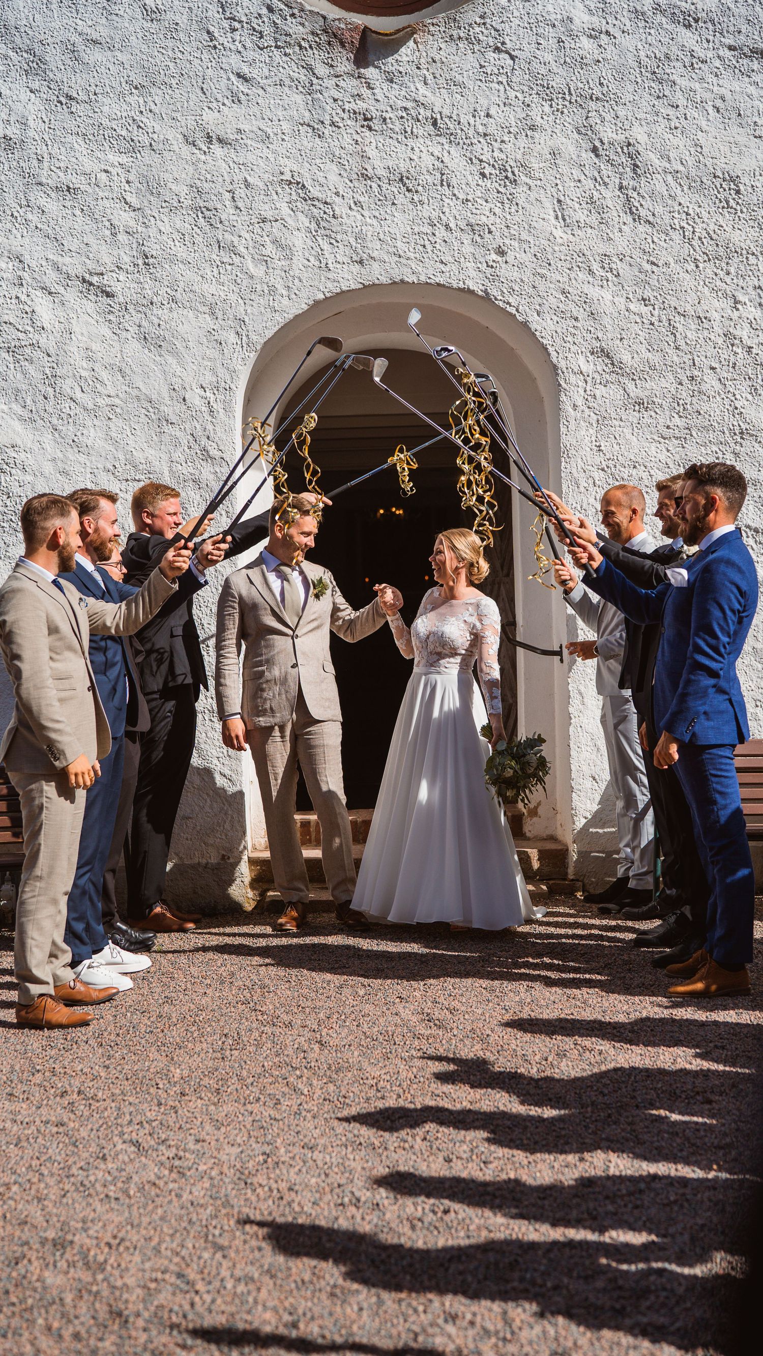Wedding party forms golf club arch as bride and groom exit white stone church after ceremony.