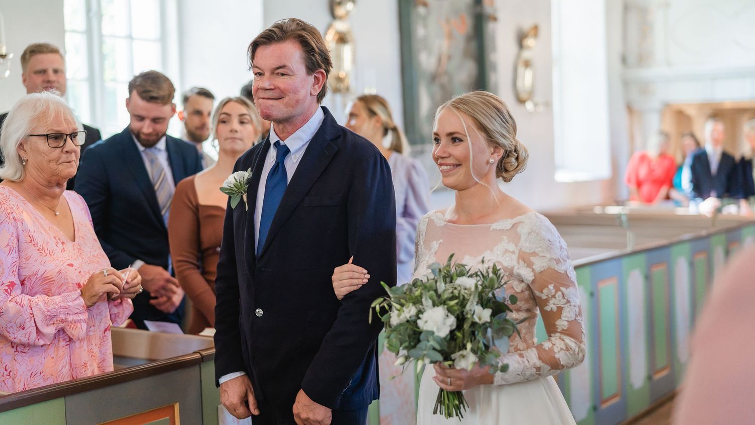 Smiling bride in white gown holding bouquet walks arm-in-arm with groom in navy suit at church wedding.