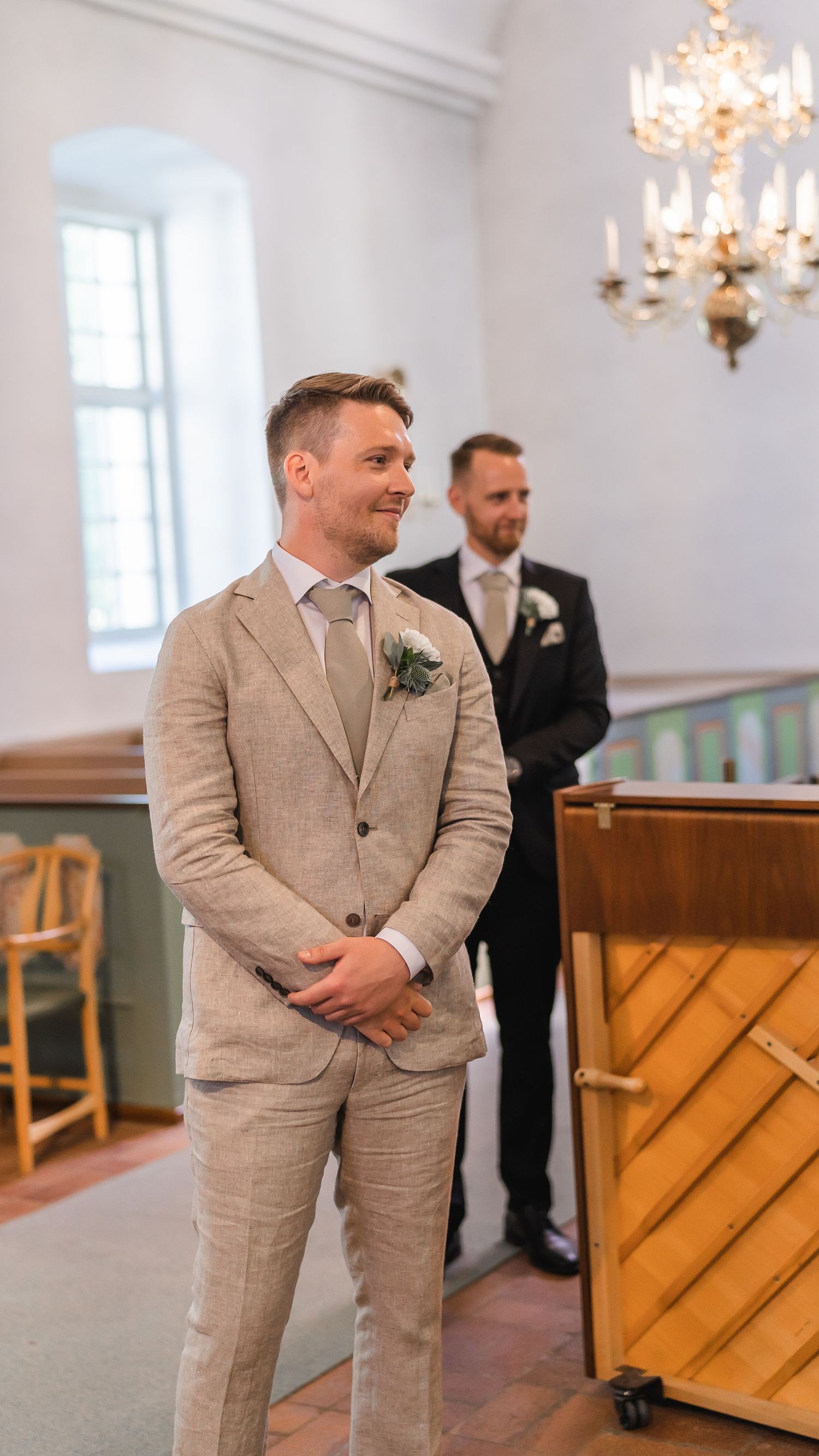Handsome groom in beige linen suit with boutonniere smiling at altar in historic church.