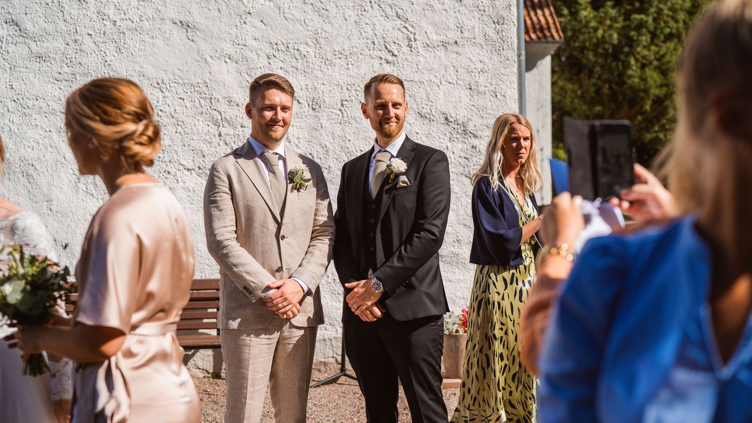 Two groomsmen in suits smiling at wedding ceremony with guests photographing in foreground.
