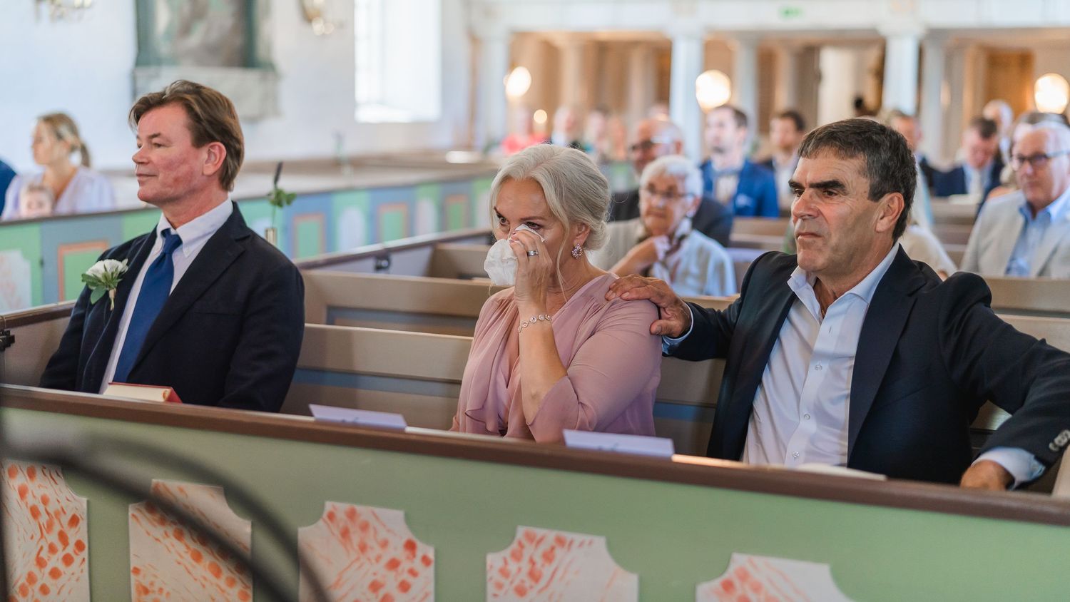 Emotional wedding guests sitting in church pews during a ceremony, with a man pointing and woman covering her mouth.