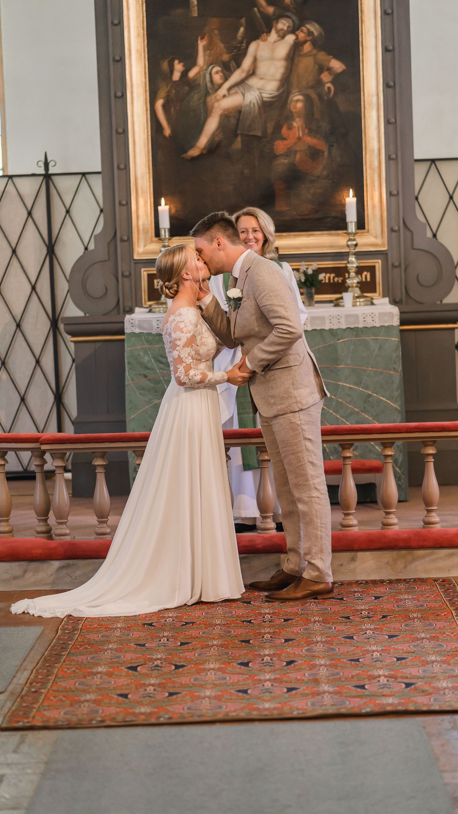 Bride and groom share their first kiss at the altar as smiling officiant watches during church wedding.