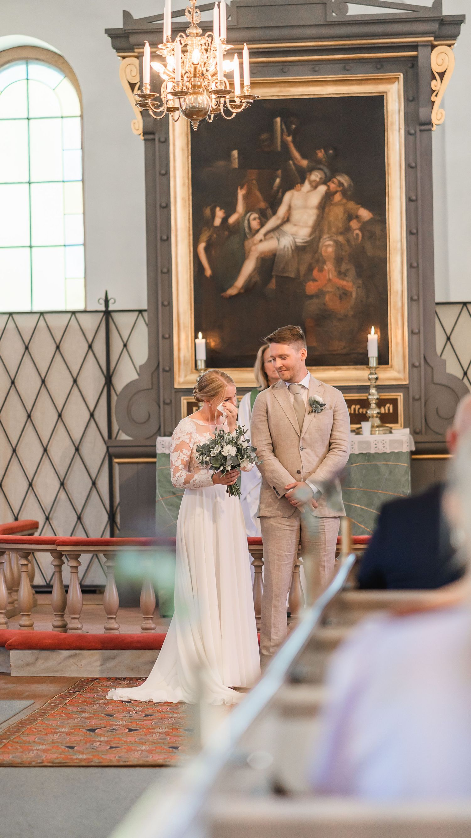 Bride in lace wedding dress smiles holding bouquet beside groom in tan suit at church altar.