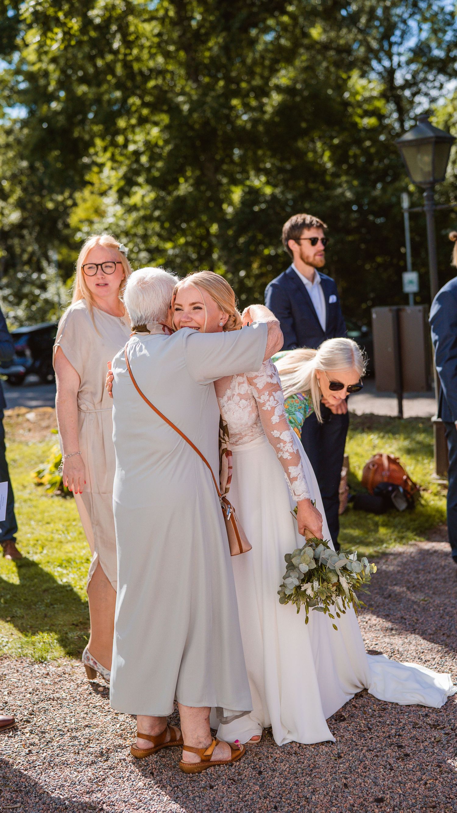 Bride in lace wedding gown embraces elderly guest outdoors while holding white floral bouquet after ceremony.