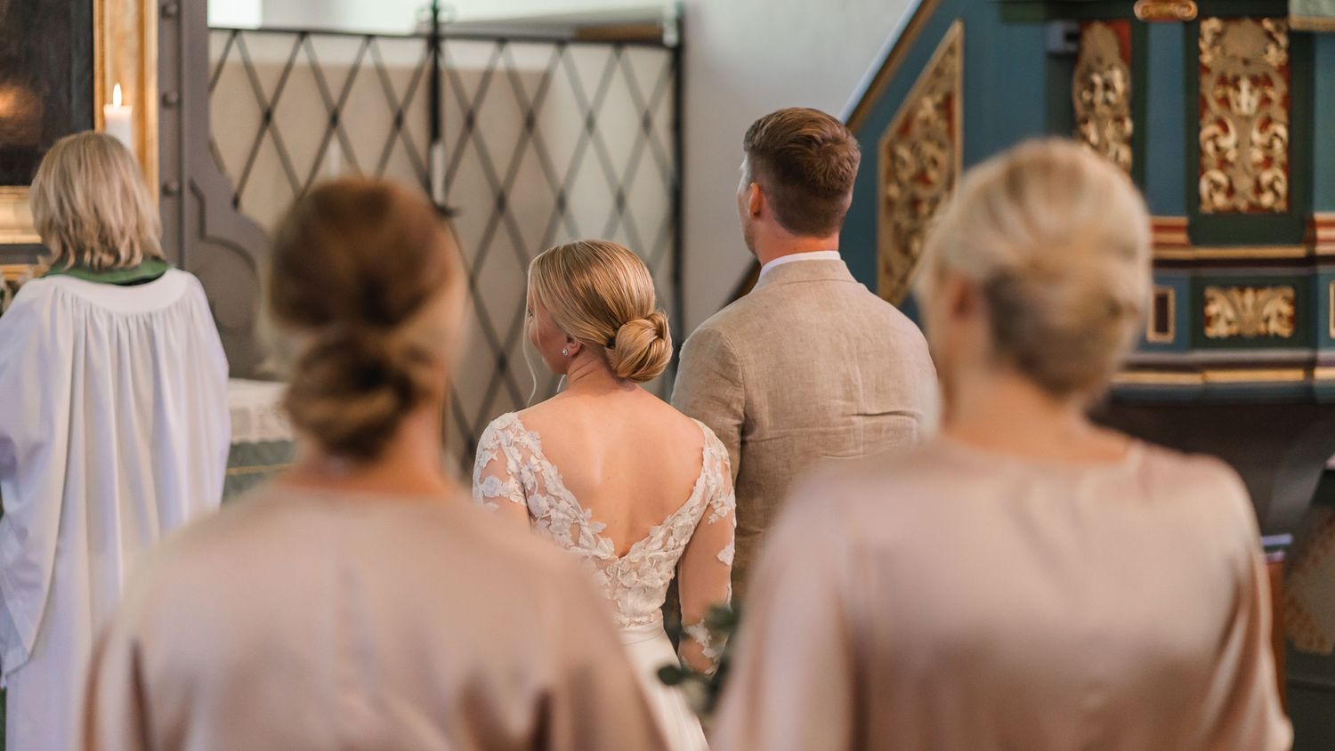 Bride with lace back wedding gown stands at altar with groom and bridesmaids during church ceremony.