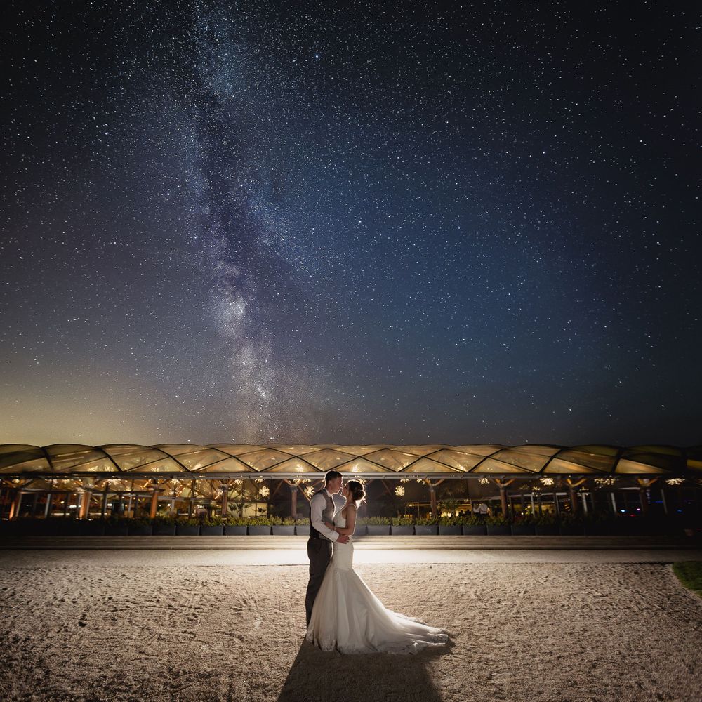Couple poses against stunning Milky Way galaxy backdrop on sandy beach at night.