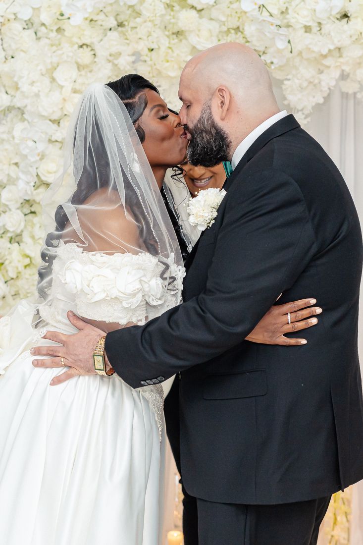 Romantic kiss between newlyweds in front of white floral wall during wedding ceremony.