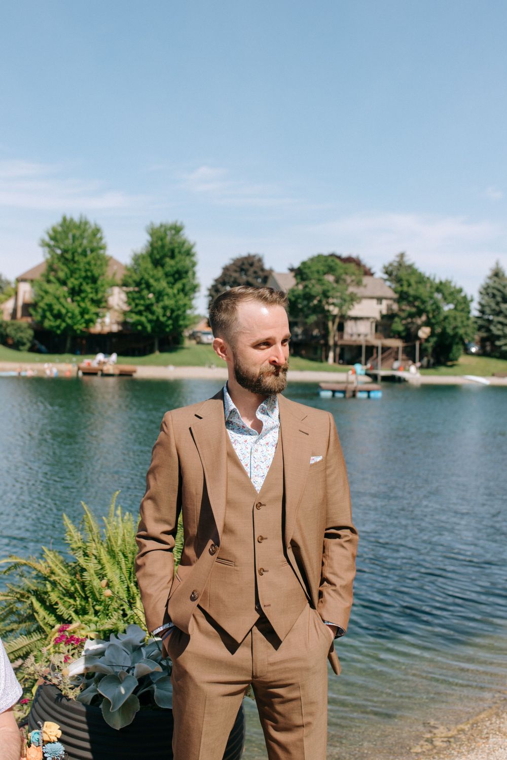 A groom in a brown suit stands by the lake during an outdoor wedding ceremony.