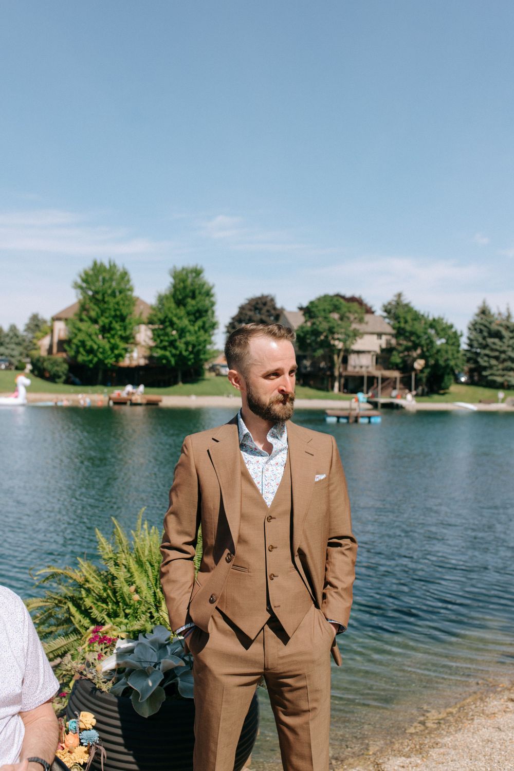 A person in a brown three-piece suit stands by a lakeside with residential homes in the background.