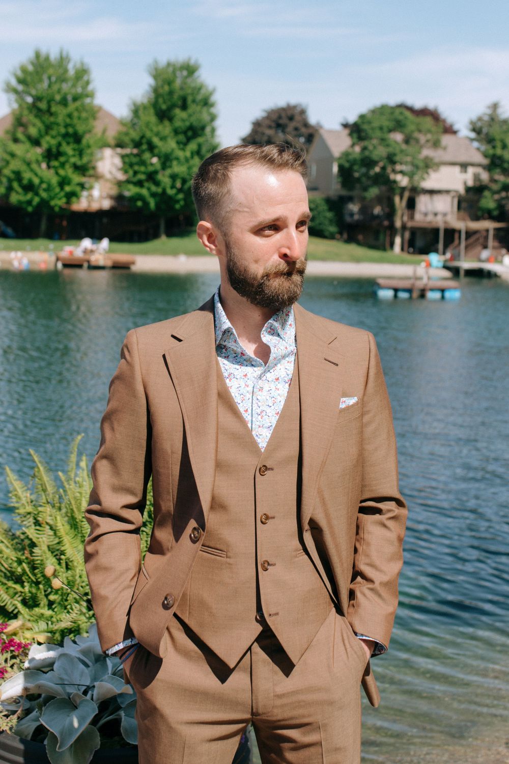 A groom wearing a tailored brown three-piece suit stands beside decorative plants on a lakefront dock.