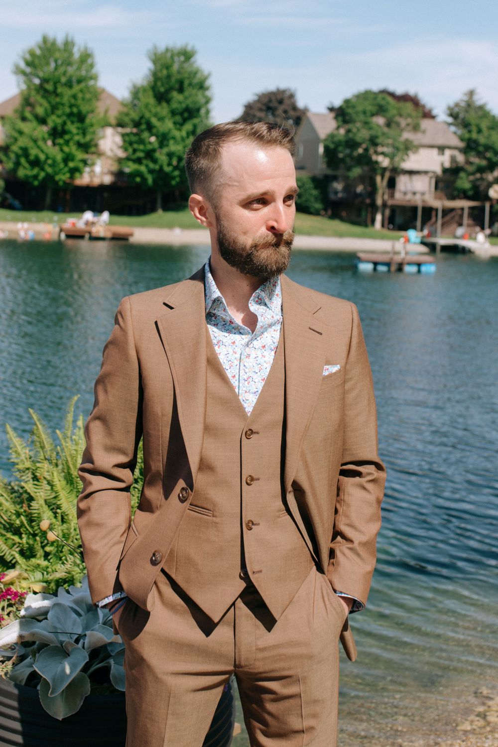 A person in a stylish tan suit poses beside a lake on a sunny day.