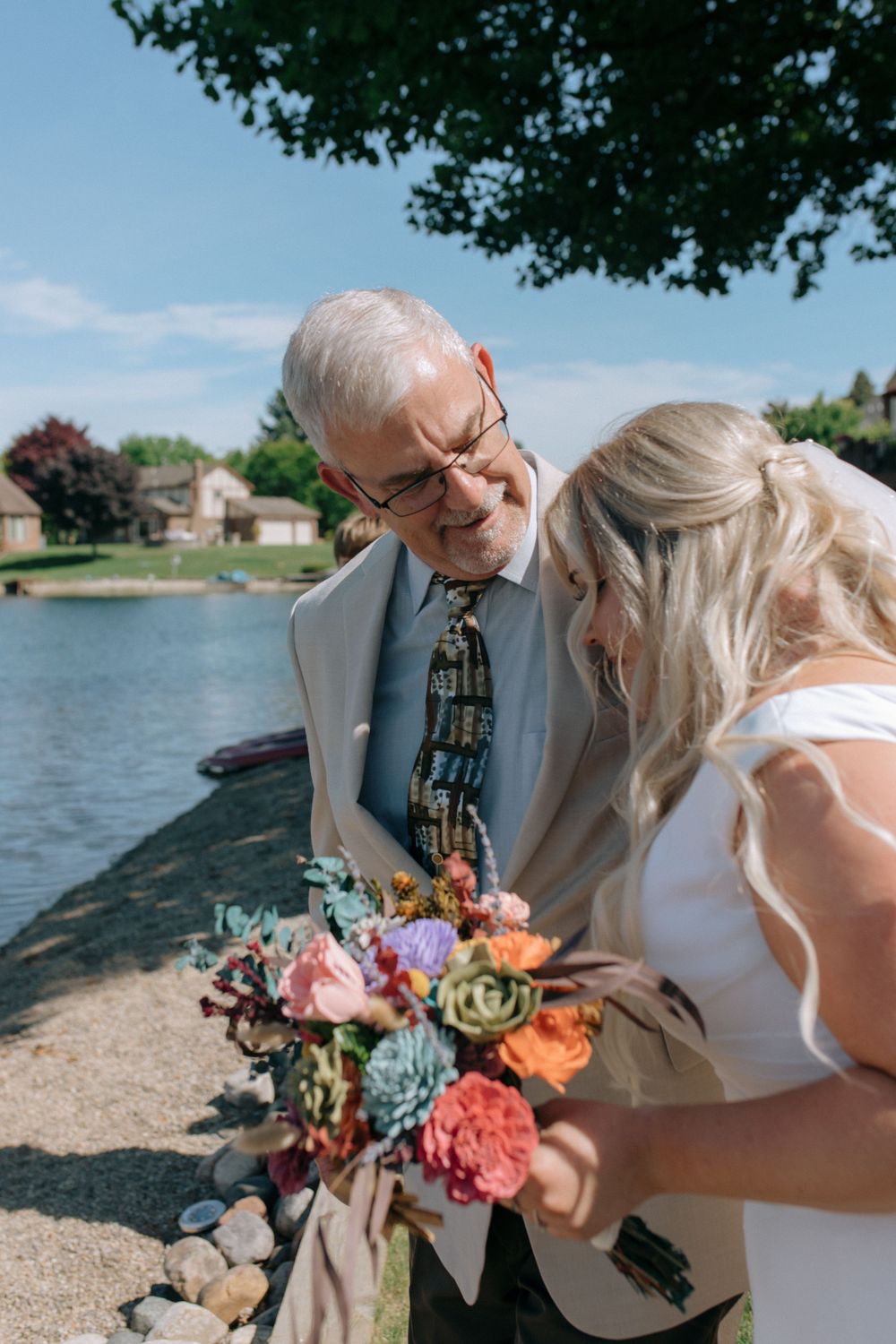 Tender moment between the bride and an older gentleman in a suit by a lakeside with a colorful bouquet.