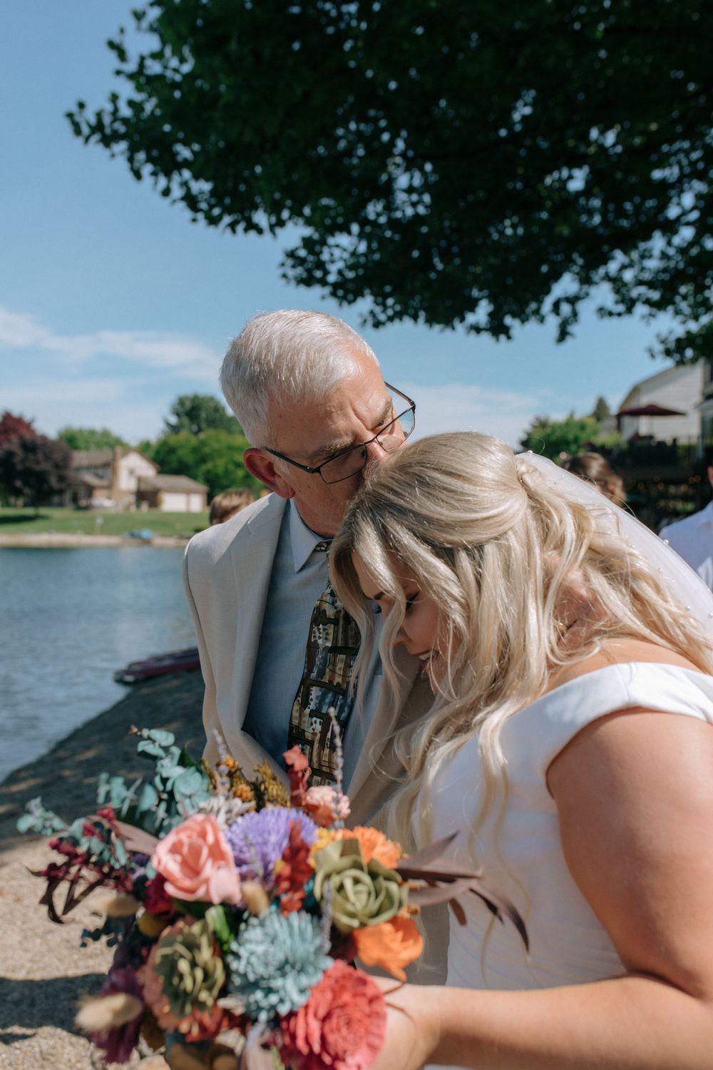 Intimate lakeside scene with bride and elderly man sharing a touching moment over a vibrant bouquet.