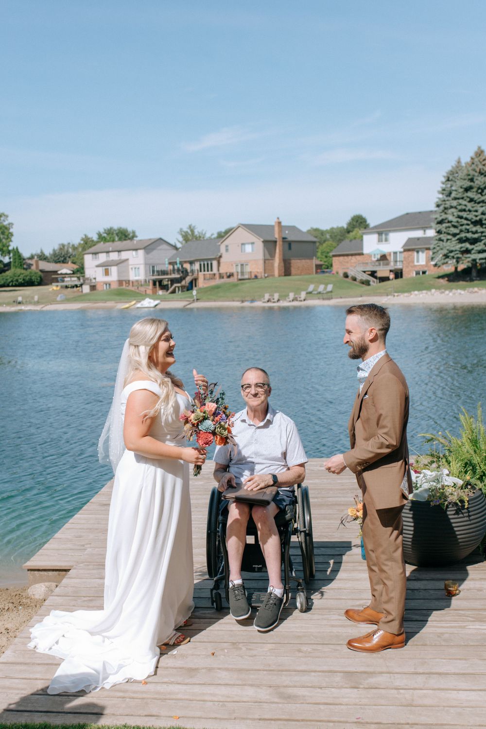 Waterfront wedding ceremony with couple and officiant in wheelchair on wooden dock.