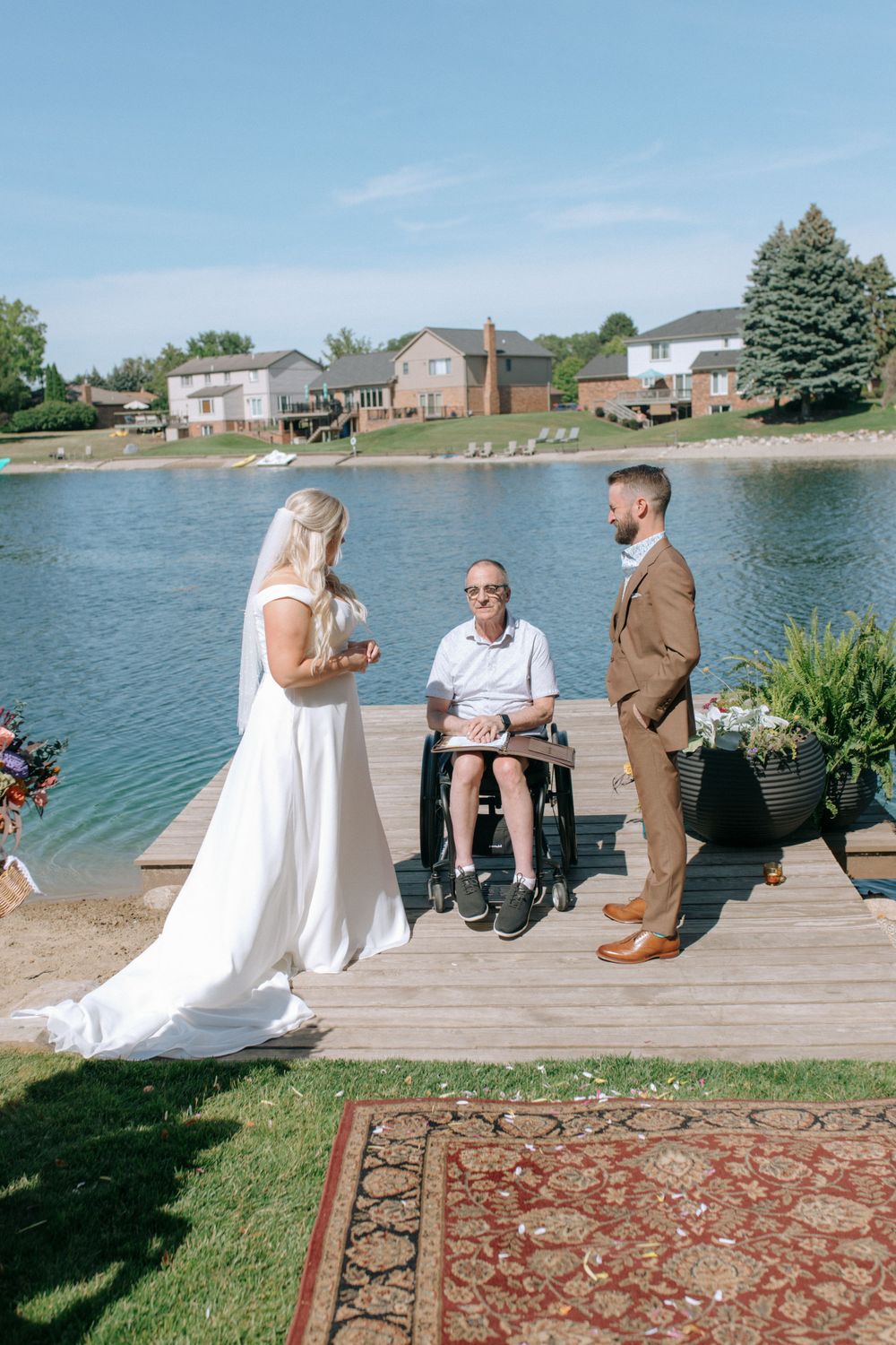 Lakeside wedding ceremony with bride, groom, and officiant in wheelchair on wooden pier.