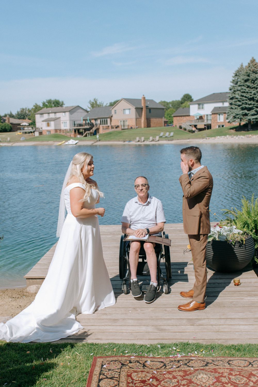 Wedding ceremony taking place on lakefront dock with guests watching from shore.