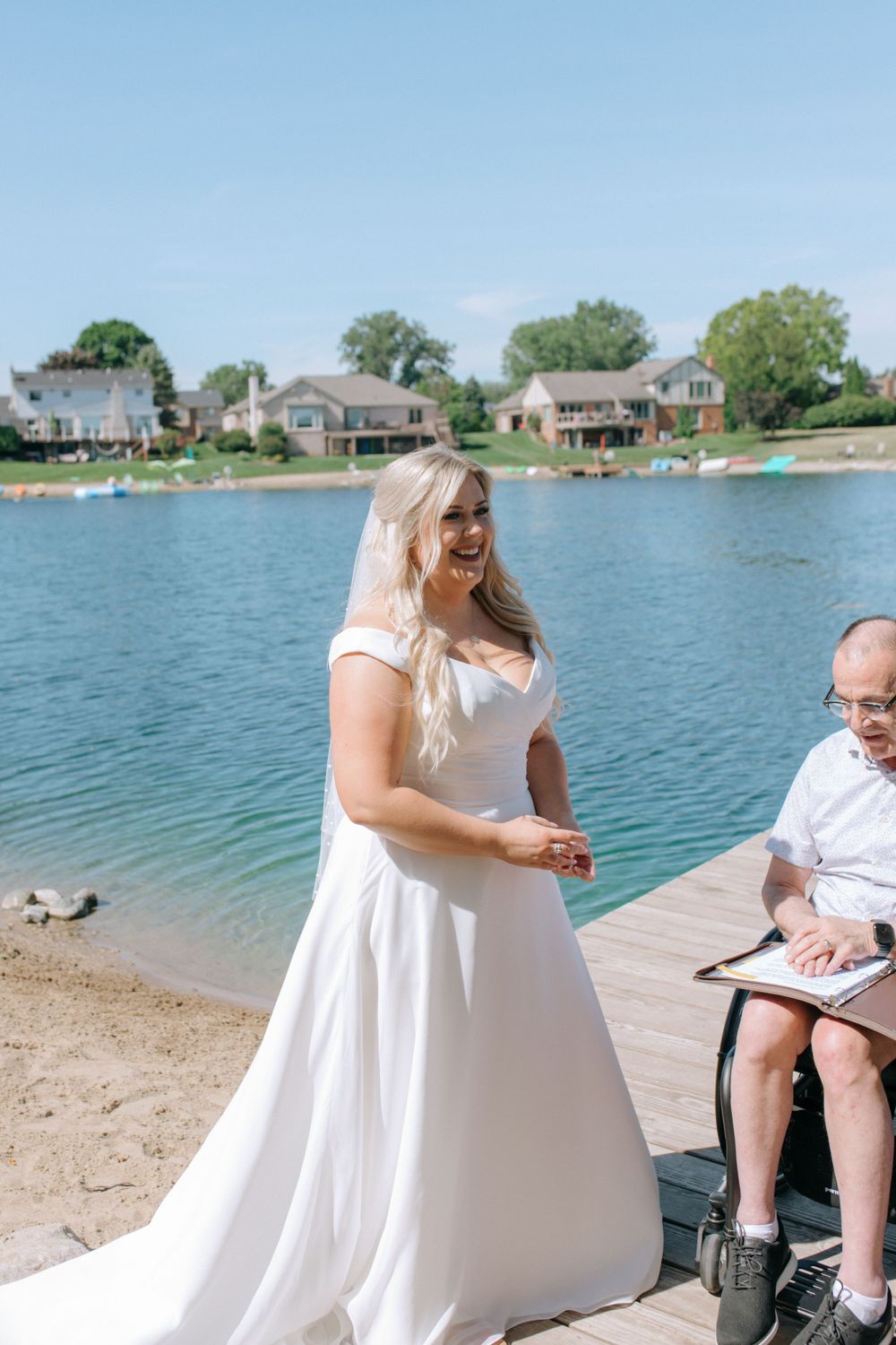 Radiant bride in white dress smiling during lakeside ceremony with officiant.