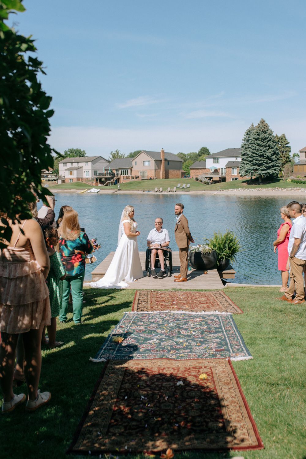 Outdoor wedding ceremony by the lake with decorative rugs and guests watching from the lawn.