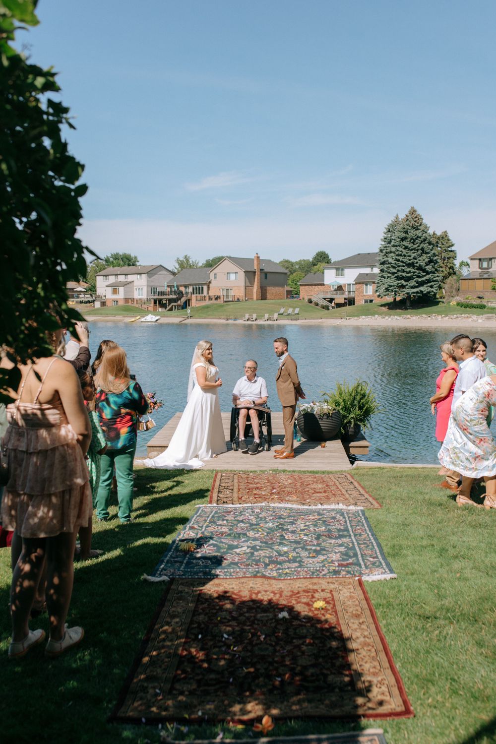Wedding guests gather on lawn to witness lakeside ceremony on decorated dock.
