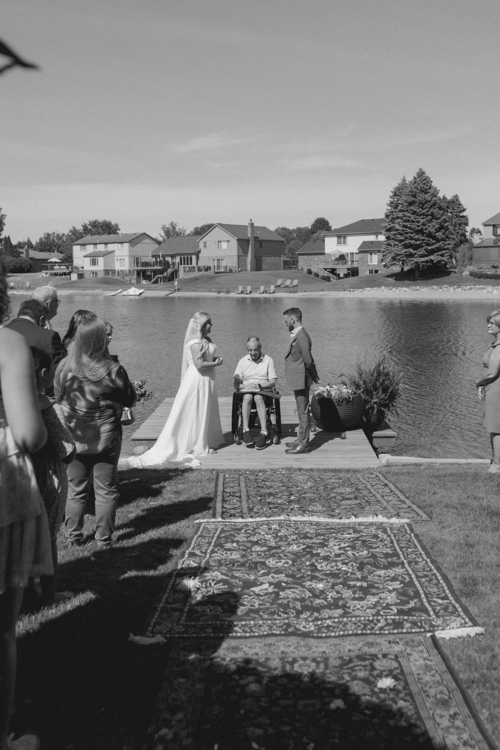 Black and white photo of waterfront wedding ceremony with guests watching from grassy shore.