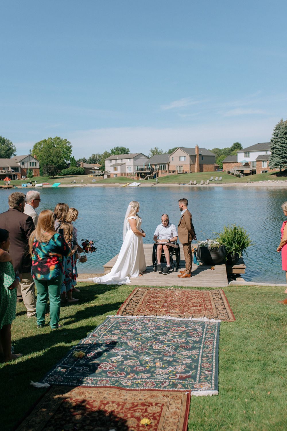 A lakeside wedding ceremony setup with ornate rugs leading to a small dock where guests gather on a sunny day.