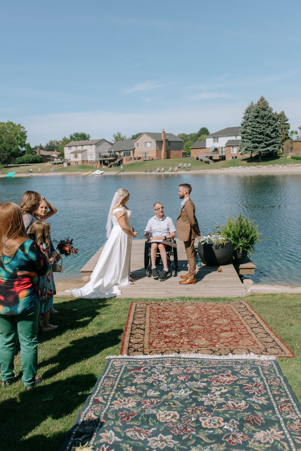 Outdoor wedding ceremony taking place on a dock overlooking a lake with decorative rugs laid on the grass.