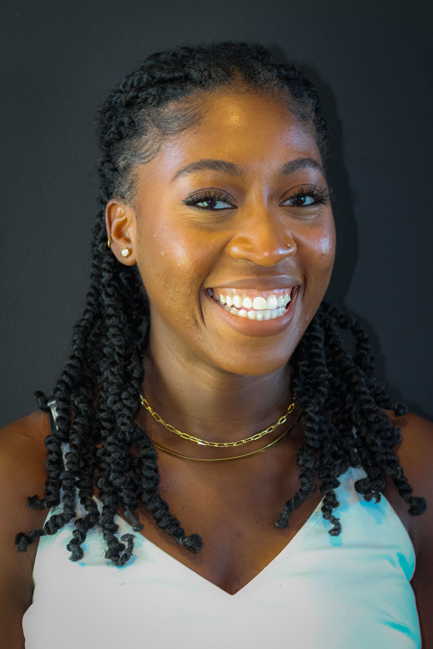 Studio portrait of someone in a white tank top with braided hair and gold necklace against dark background.