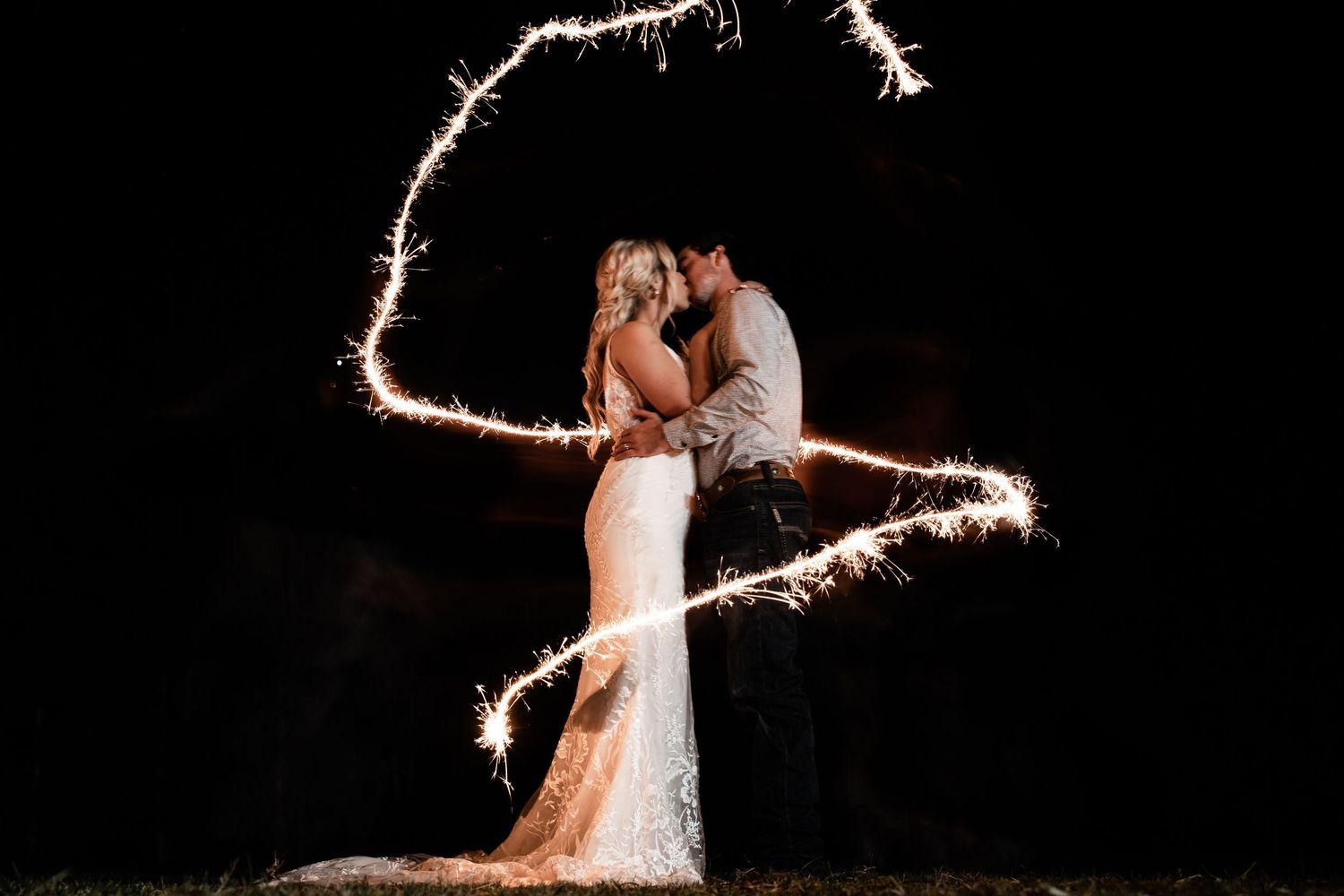 Romantic wedding photo at night with sparklers creating a heart shape around a couple in an elegant gown and suit.