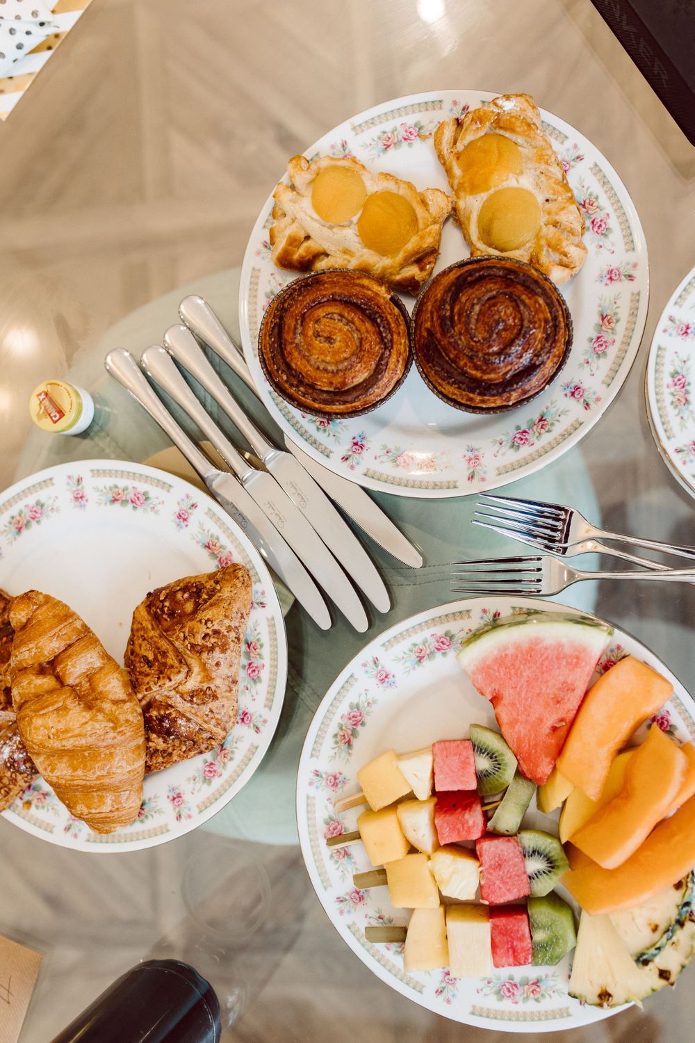 A breakfast spread on floral plates with pastries, watermelon, and fruit served with plastic utensils.