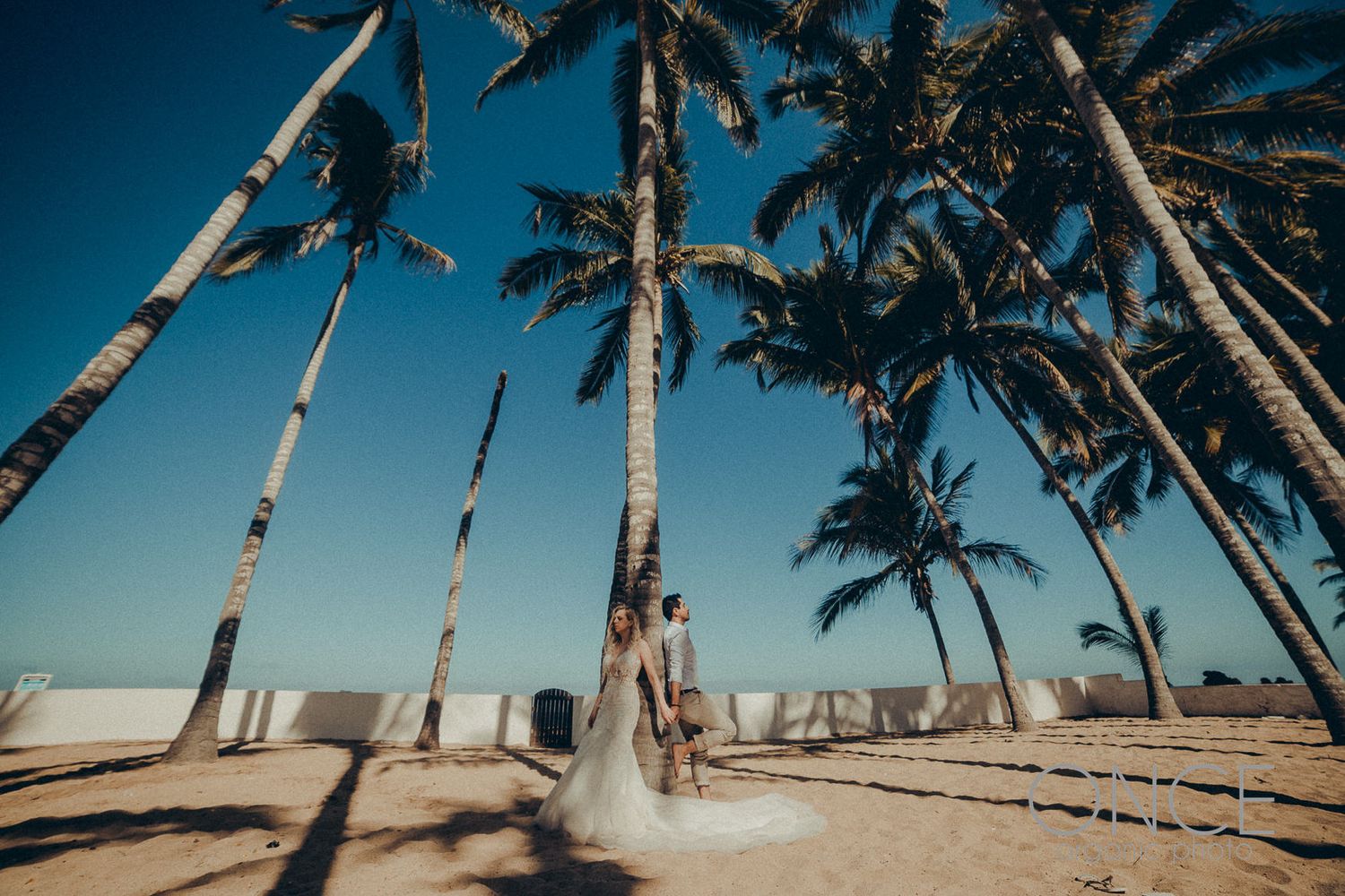 A wedding dress flows in the wind among tall palm trees on a tropical beach.