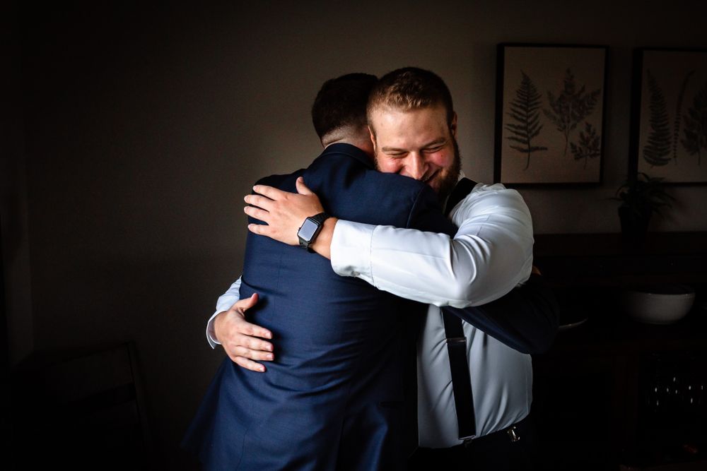 Emotional embrace between wedding party members wearing navy suits in dimly lit room.
