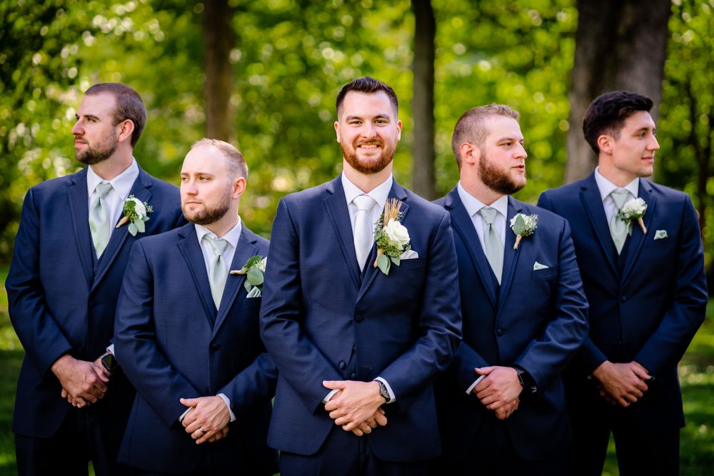 Groomsmen in navy suits lined up outdoors with white boutonnieres against a green natural background.