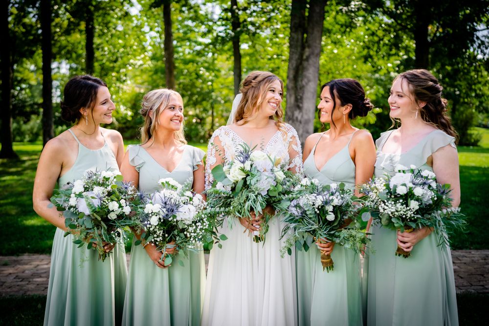 Bridesmaids in sage green dresses holding white floral bouquets pose together outdoors.