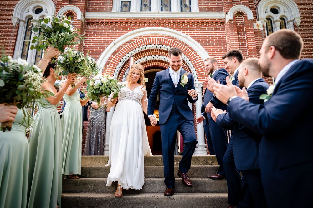 Wedding party members jump and pose playfully in front of a red brick church with white archways.