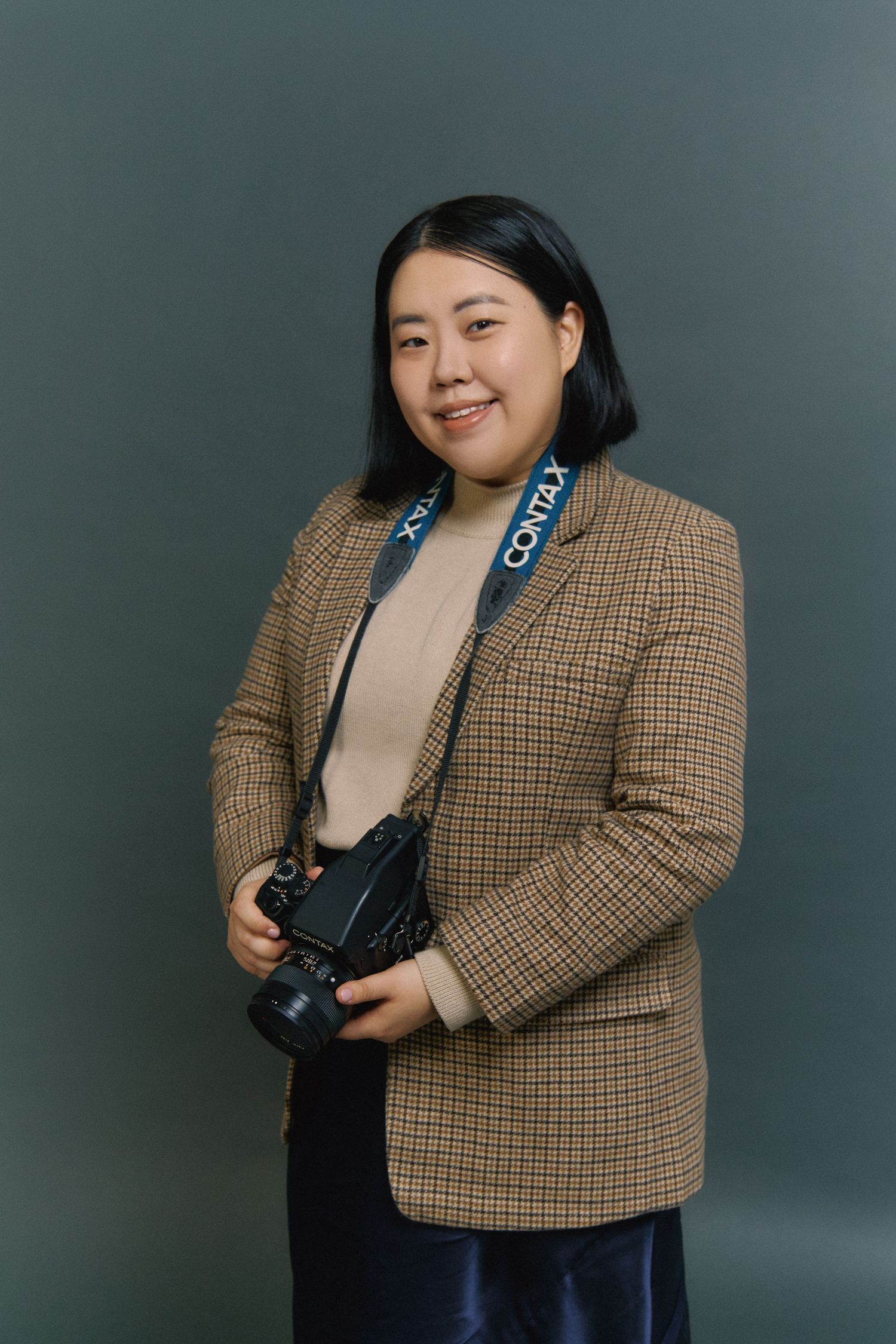 Professional photographer wearing a checkered blazer and holding a camera against a gray backdrop.